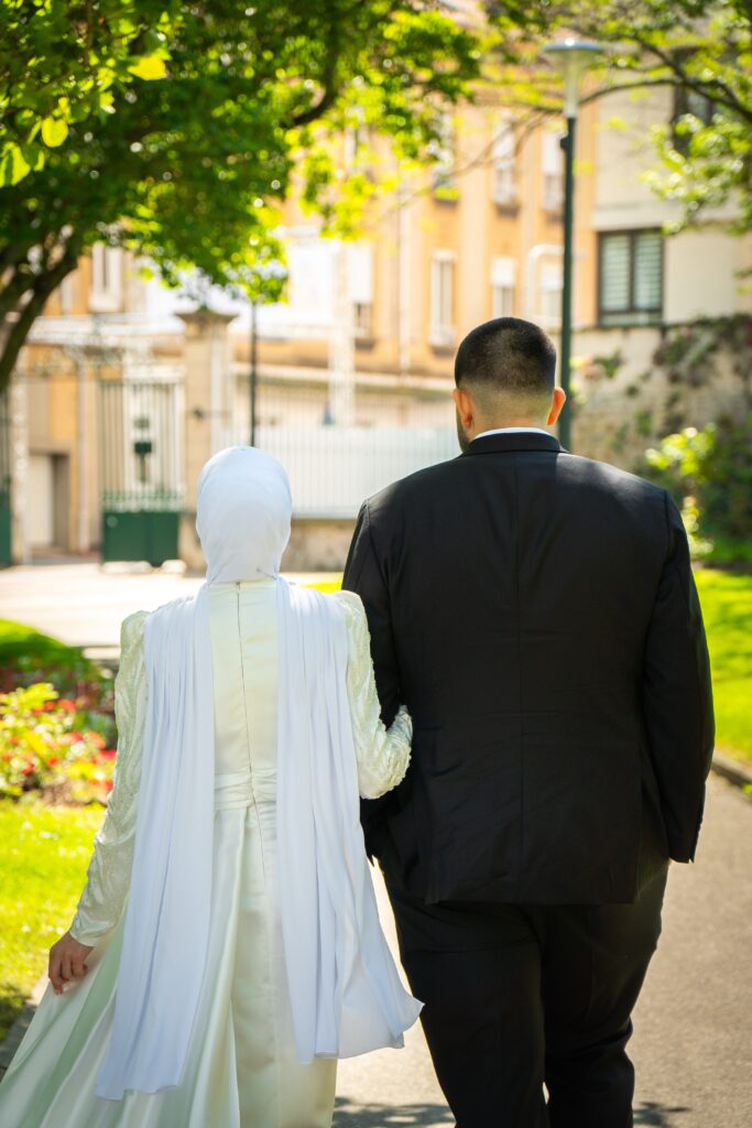 Deux mariés de dos, marchant main dans la main dans un parc à Saint-Chamond, entourés de verdure. Capture romantique d’un moment intime lors de leur mariage.